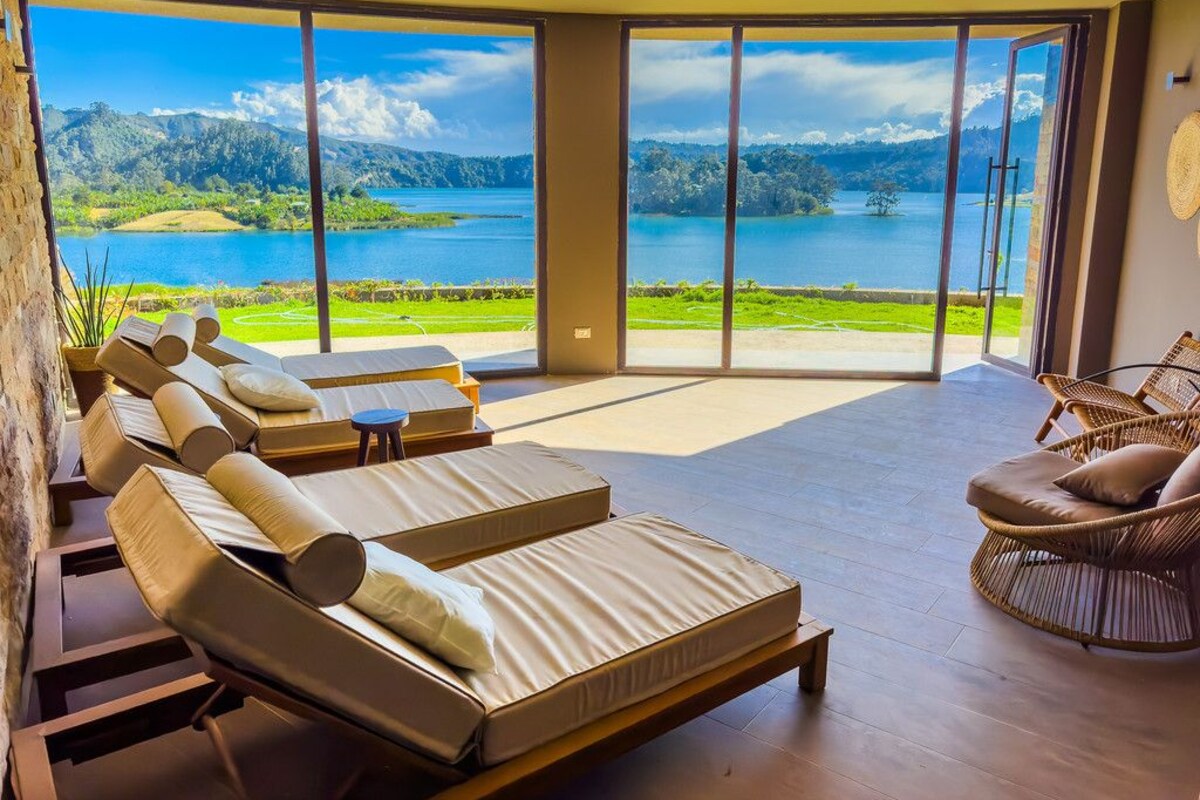 A sunlit room with large windows overlooking a lake and mountains, furnished with lounge chairs and a wicker chair.