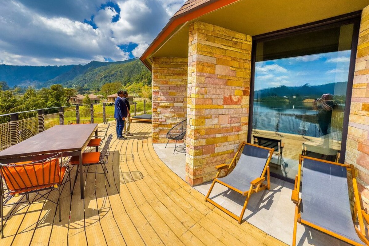 A wooden deck with a table and chairs, a glass-enclosed area with a view of mountains, and a person standing by the railing.
