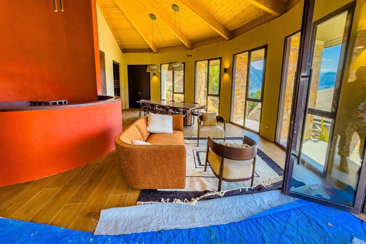 A spacious room with a wooden ceiling, large windows, a curved sofa, a dining table, and a fireplace.