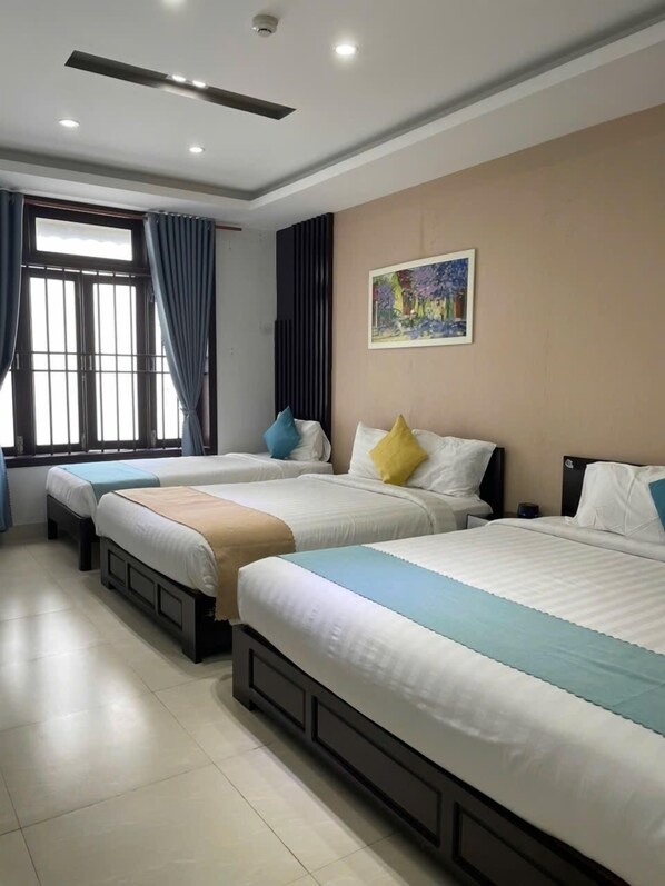 Habitación de hotel con tres camas, un ventanal con cortinas, ventilador de techo, iluminación empotrada y un cuadro en la pared.