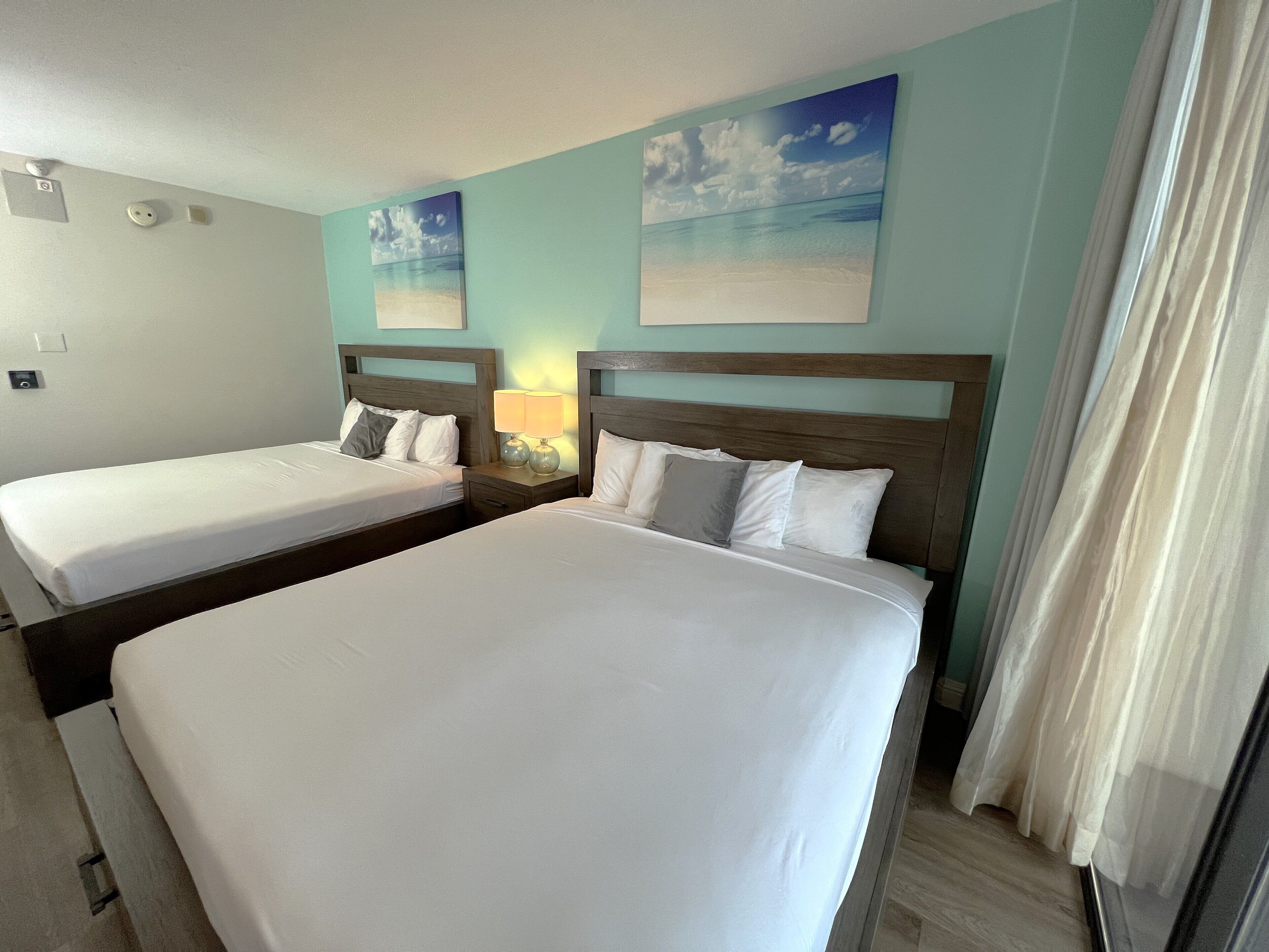 A hotel room with two single beds, a nightstand with a lamp, and a painting of a beach scene on the wall.