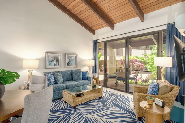 A modern living room with a blue and white patterned rug, a glass coffee table, a sofa, and a wicker chair. There are two wall-mounted lamps and a large window with a view of greenery outside.