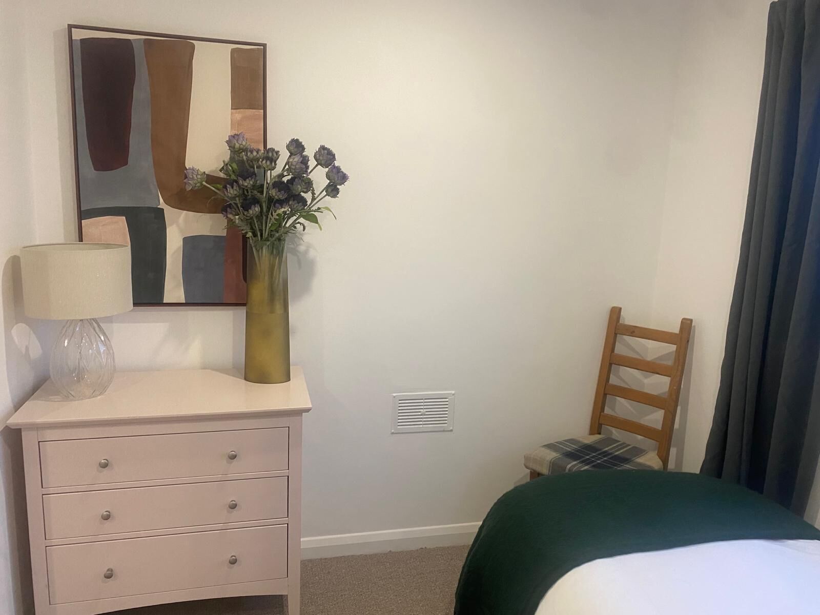 A bedroom with a bed, a bedside table with a lamp, a vase with flowers, a chair, and a painting on the wall.
