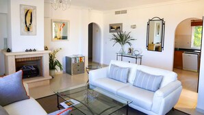 Un salon moderne avec un canapé blanc, une table basse en verre, une cheminée et une vue sur la cuisine.