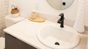 A bathroom with a white sink, a round mirror, a towel rack, and a small wooden stool. - The Georgie Studio/1Bath Located in the heart of Downtown (Sitka)