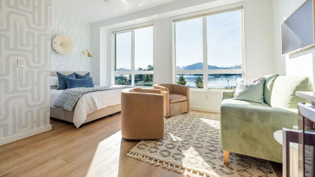 A modern bedroom with a bed, two armchairs, a green sofa, and large windows overlooking a body of water. - The Georgie Studio/1Bath Located in the heart of Downtown (Sitka)