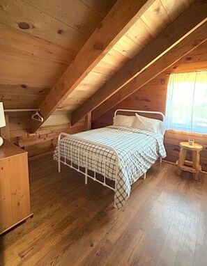 A wooden cabin bedroom with a bed, a nightstand, a small stool, and a window with curtains. - Cozy 3 bedroom, 3 bathroom, Log Home, PET FRIENDLY, Tollgate (Sisters)