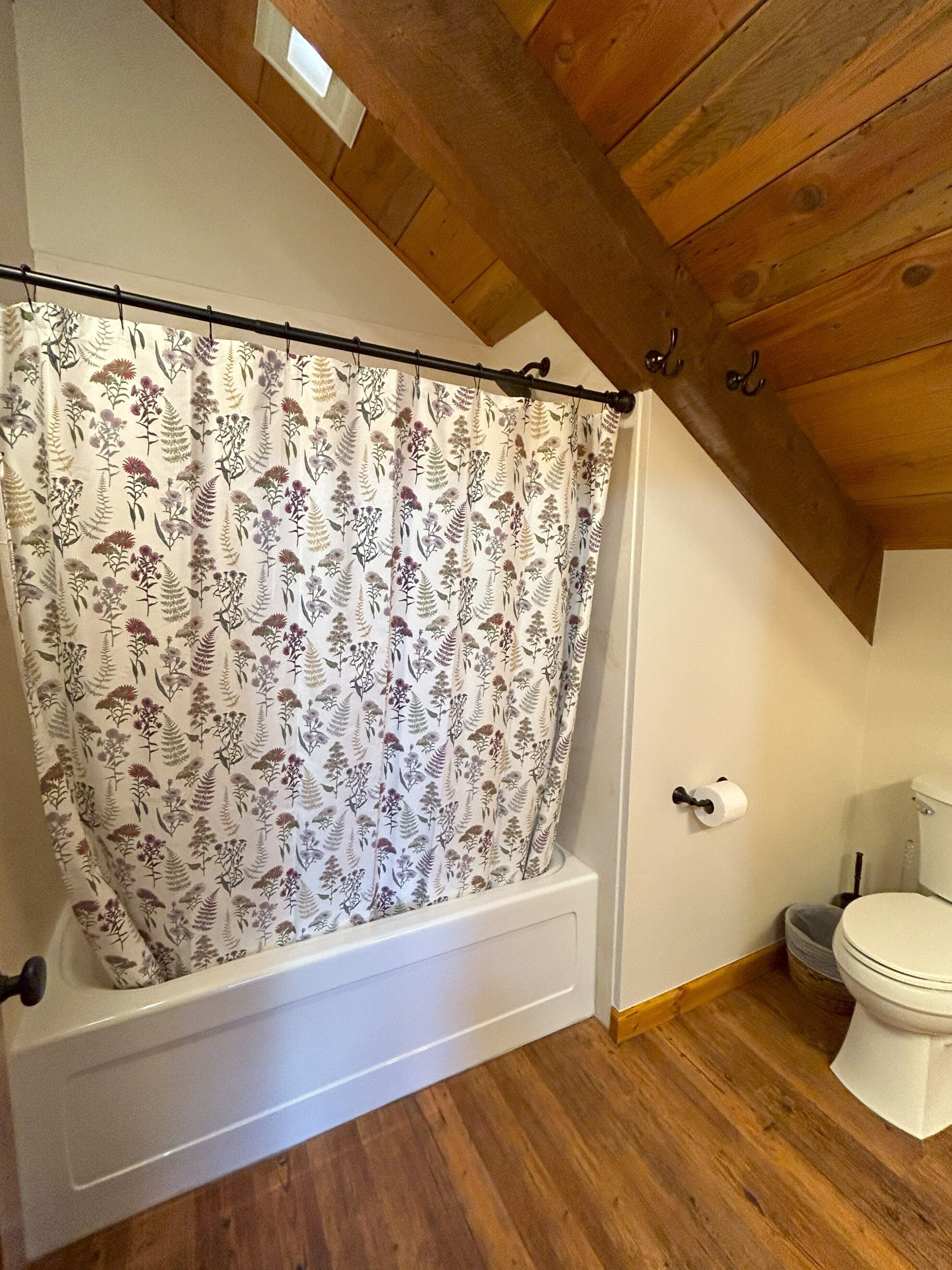 A bathroom with a shower curtain featuring a floral pattern, a white bathtub, a toilet, and wooden flooring.