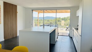 A modern kitchen with a central island, a dining area with yellow chairs, and a view of a balcony with outdoor furniture.
