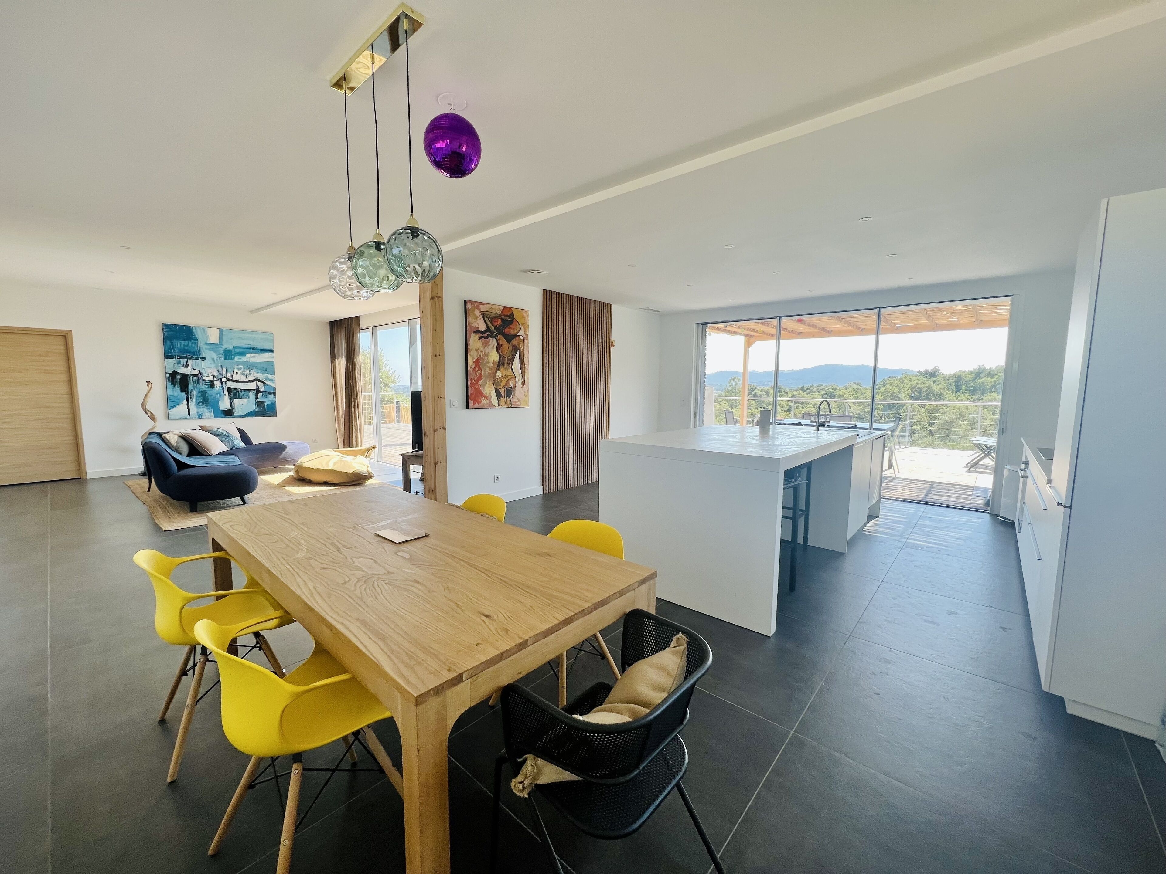 A modern kitchen with a central island, a dining area with a wooden table and yellow chairs, and a living area with a sofa and a painting.