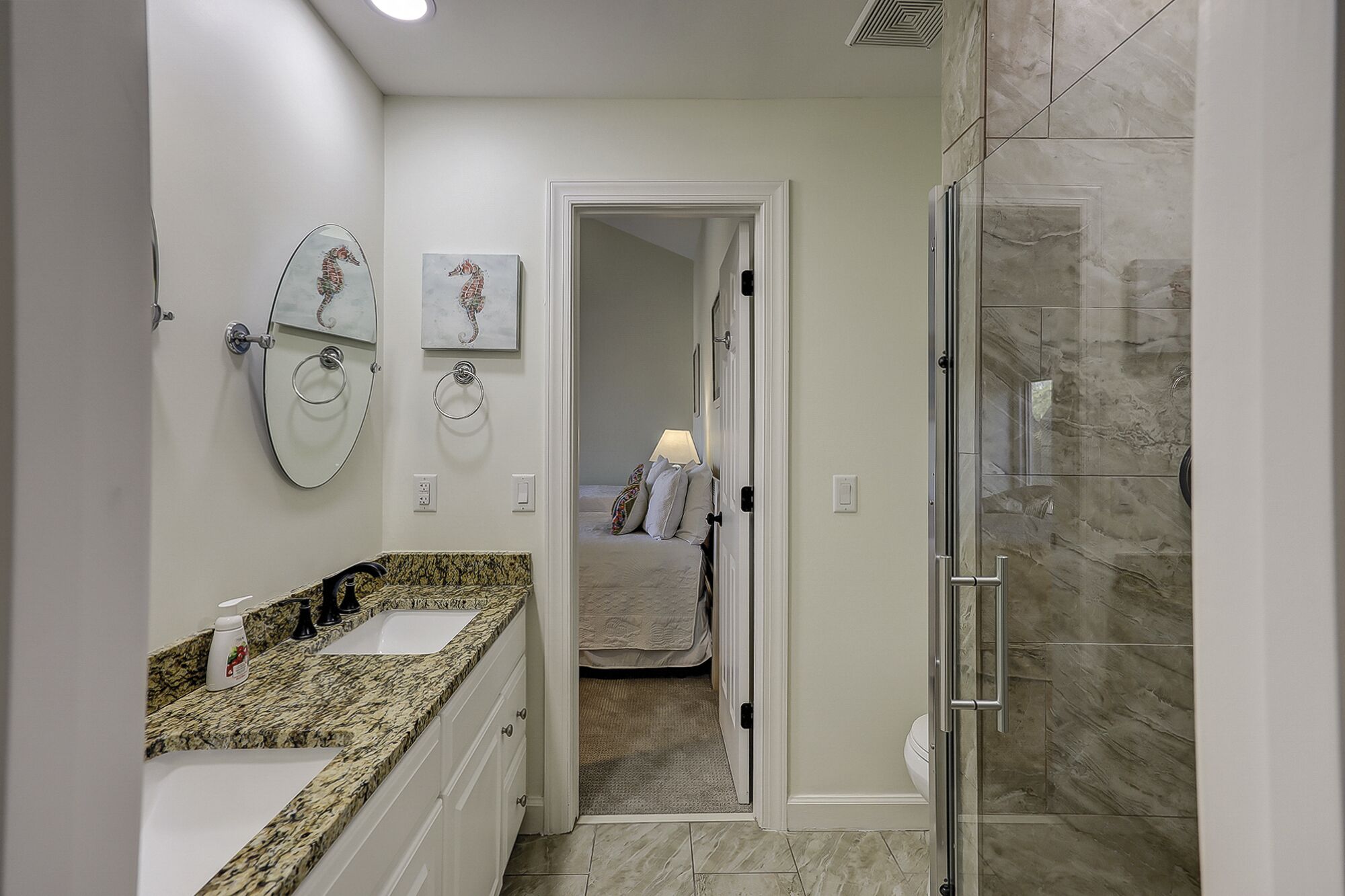 A modern bathroom with a granite countertop, a glass shower enclosure, and a bed visible through an open door.