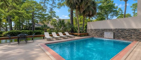 A rectangular swimming pool with a wooden deck and a stone wall, surrounded by trees and a clear sky.