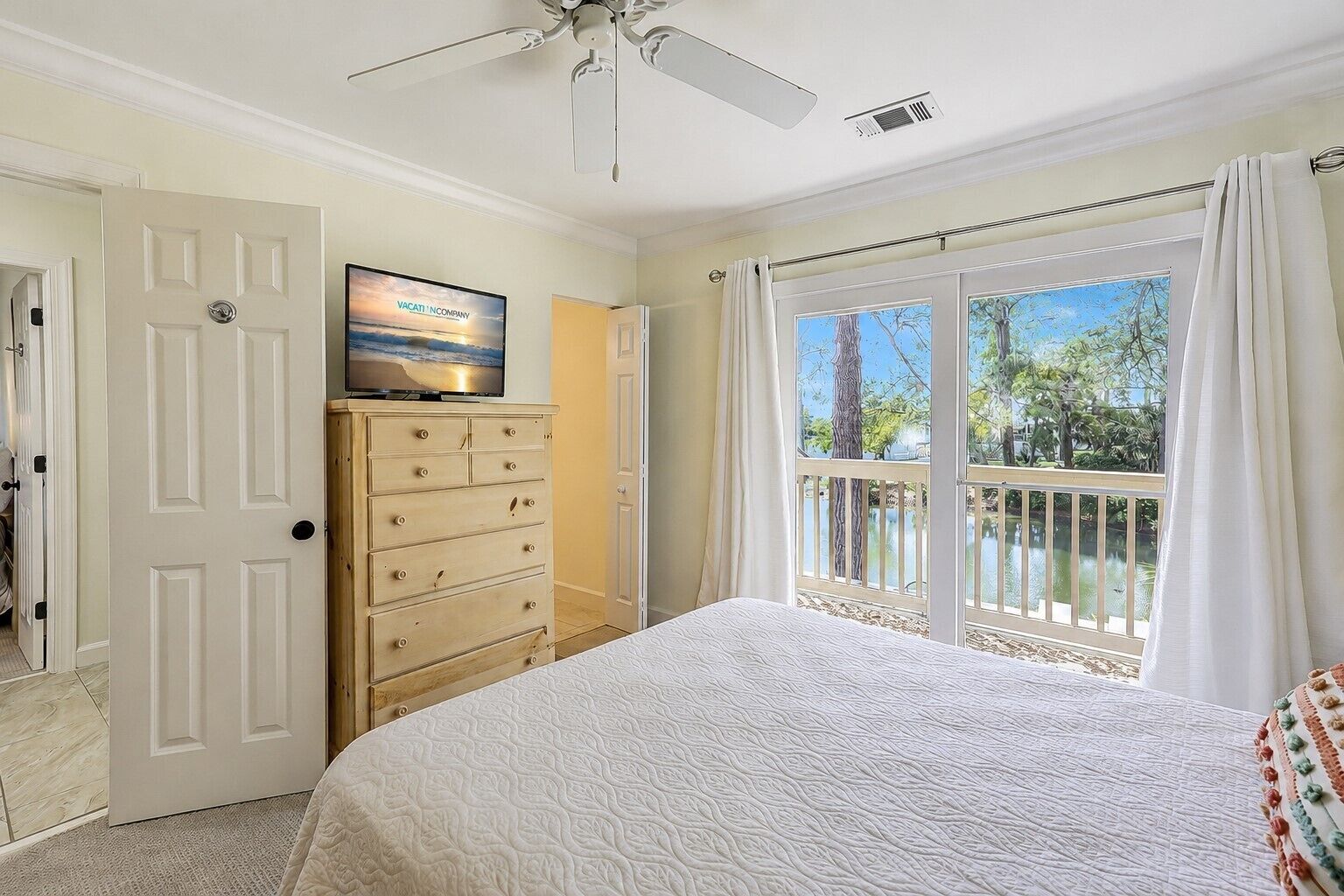 A bedroom with a bed, a dresser, a television, and a balcony with a view of trees and a body of water.