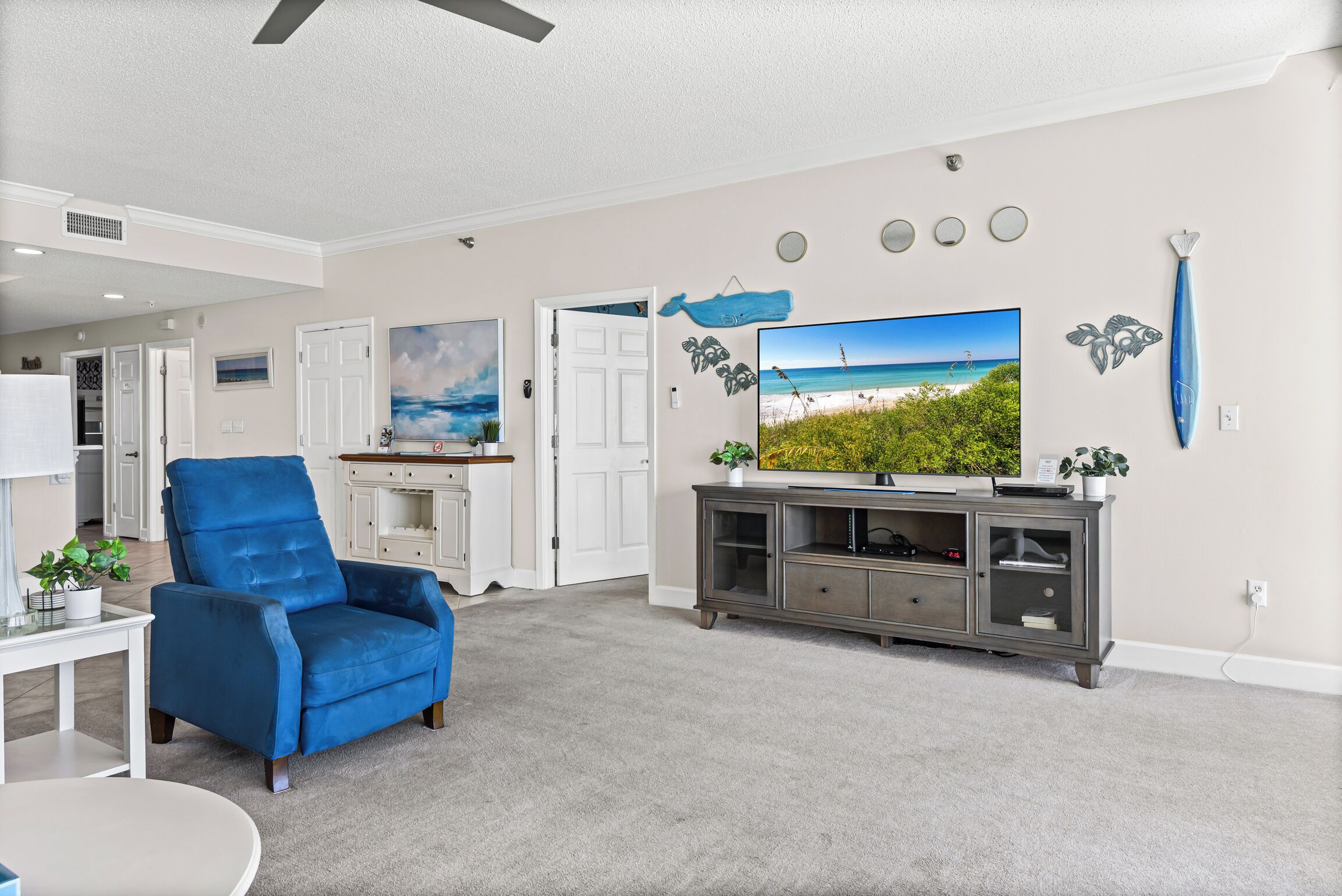 A living room with a large flat-screen TV, a blue armchair, a grey entertainment unit, and a door leading to another room.