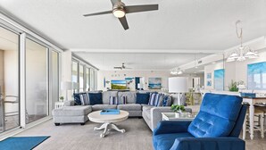 A modern living room with a grey sectional sofa, a blue armchair, and a round coffee table. There is a ceiling fan, a chandelier, and a large window providing natural light. - Gulf Front 3/3 Resort w/Pool & 2 King Suites. Chair/umbrella set-up included! (Navarre)