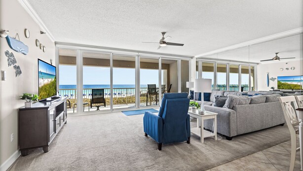 A spacious living room with a large grey sofa, a blue armchair, and a flat-screen TV. The room has a balcony with a view of the beach and ocean. - Gulf Front 3/3 Resort w/Pool & 2 King Suites. Chair/umbrella set-up included! (Navarre)