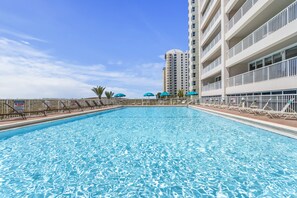 A swimming pool with lounge chairs and umbrellas, surrounded by a fence, with a tall building in the background.