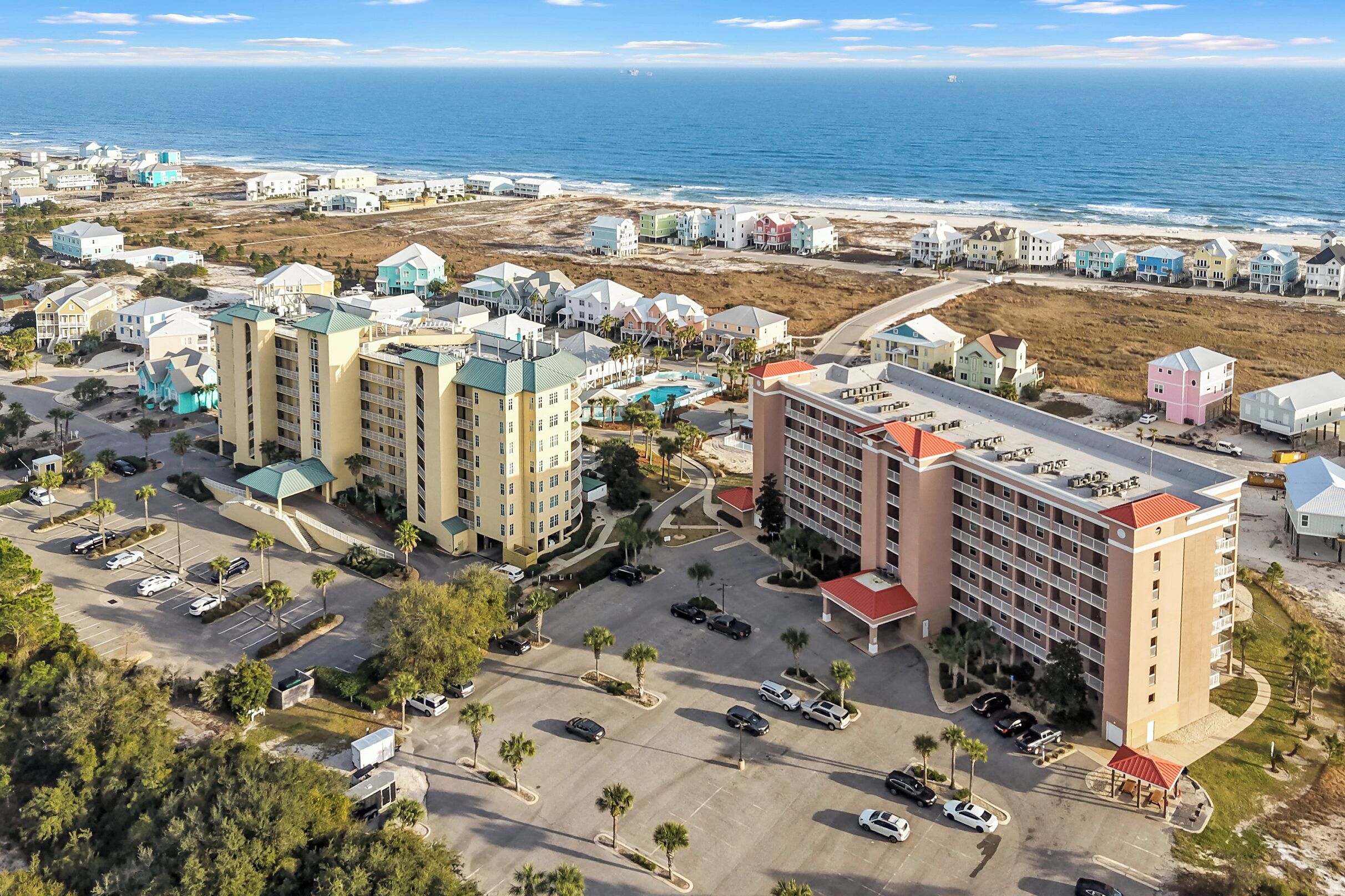 An aerial view of a coastal hotel area with multiple buildings, a parking lot, and a beach in the background.