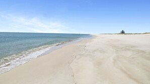 A wide sandy beach with gentle waves, a clear sky, and a small building in the distance.