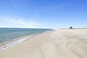A wide sandy beach with gentle waves, a clear sky, and a small building in the distance. - Ft Morgan Beach House | Pet-Friendly | Gulf View + Sleeps 16 (Gulf Shores)