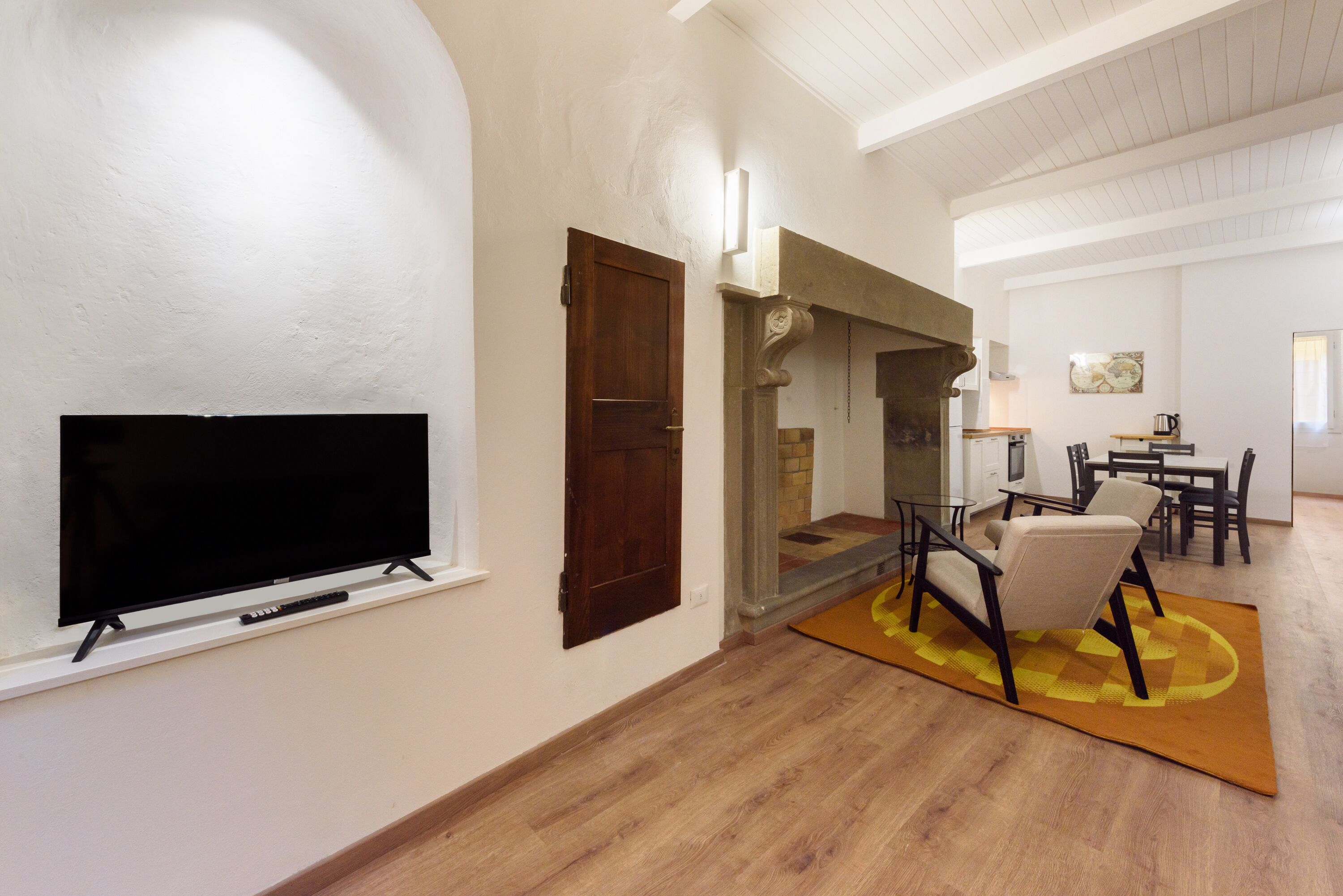 A living room with a flat-screen TV mounted on the wall, a wooden door, and a dining area with a table and chairs.