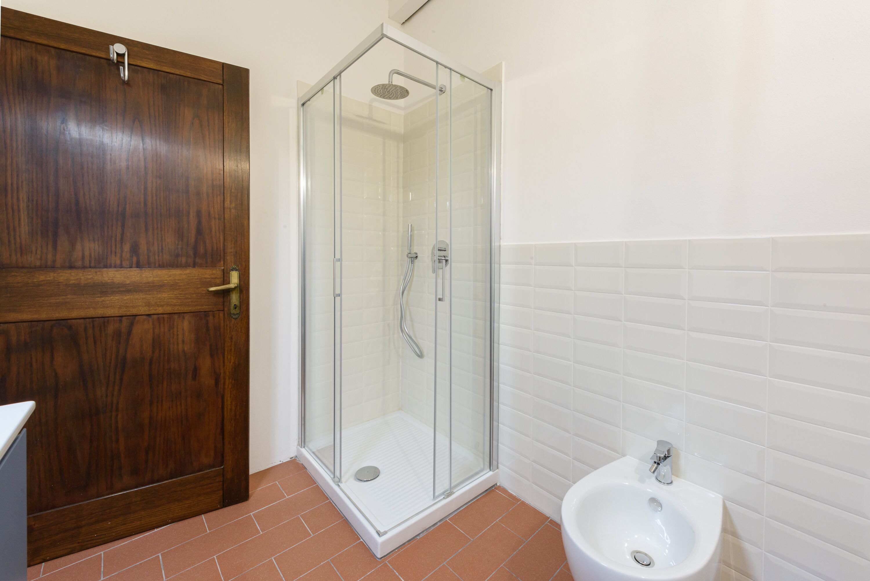 Ein Badezimmer mit Holztür, Duschkabine aus Glas und Waschbecken in Weiß.
