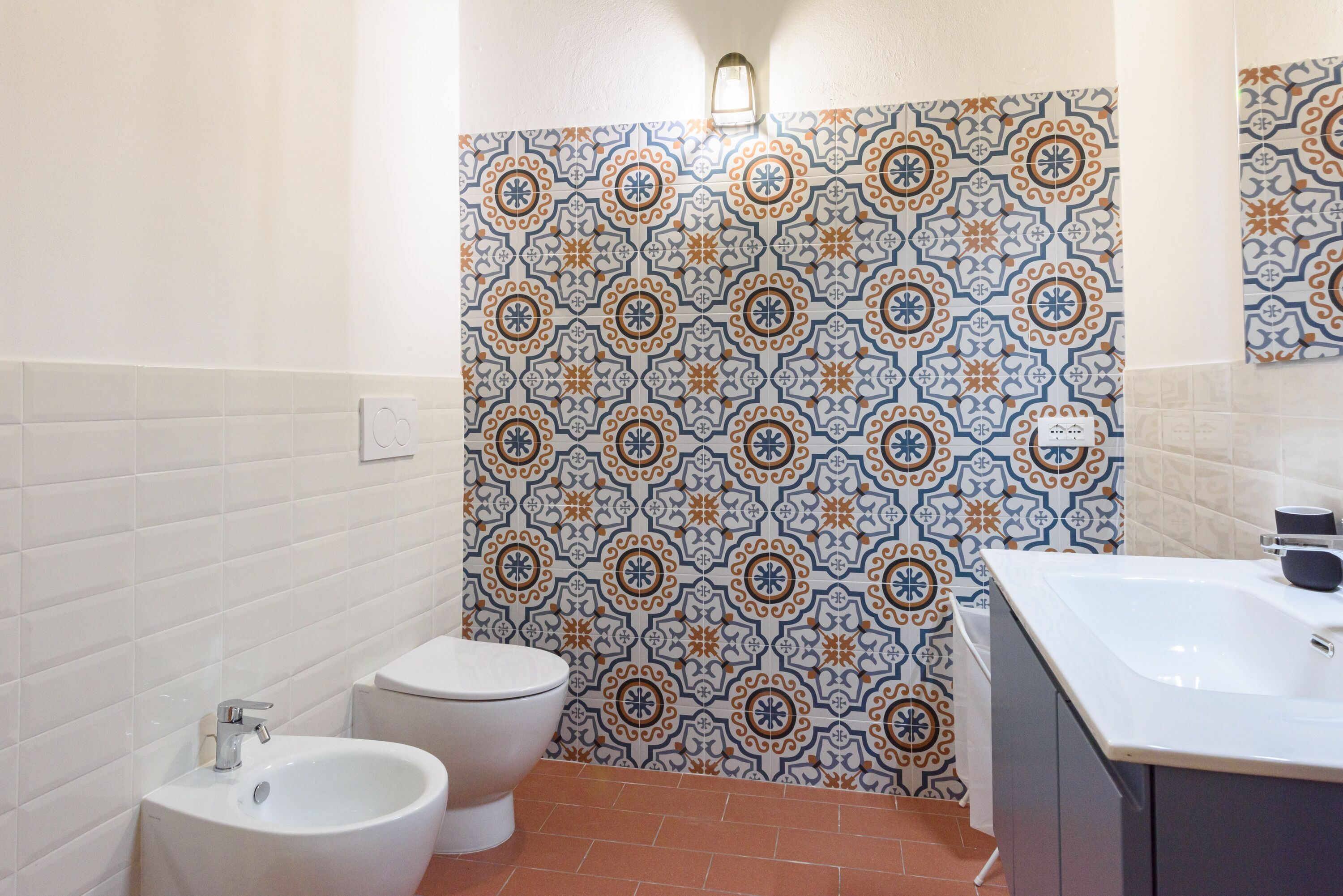 Une salle de bain avec un lavabo blanc, des toilettes et un bidet. Les murs sont recouverts de carreaux aux motifs décoratifs.