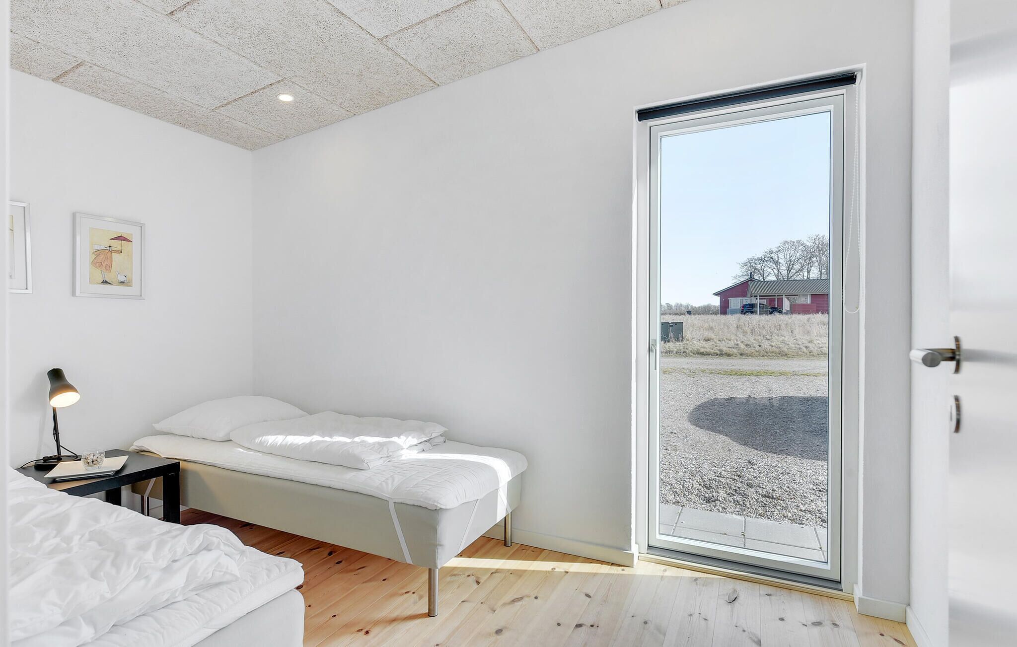 A bedroom with a bed, a desk, a lamp, and a large window with a view of a house and landscape.