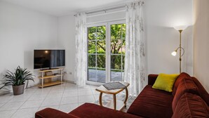 Ein Wohnzimmer mit Fernseher, einer Couch, einem Couchtisch, einer Topfpflanze und einer Glastür, die in einen Garten führt.