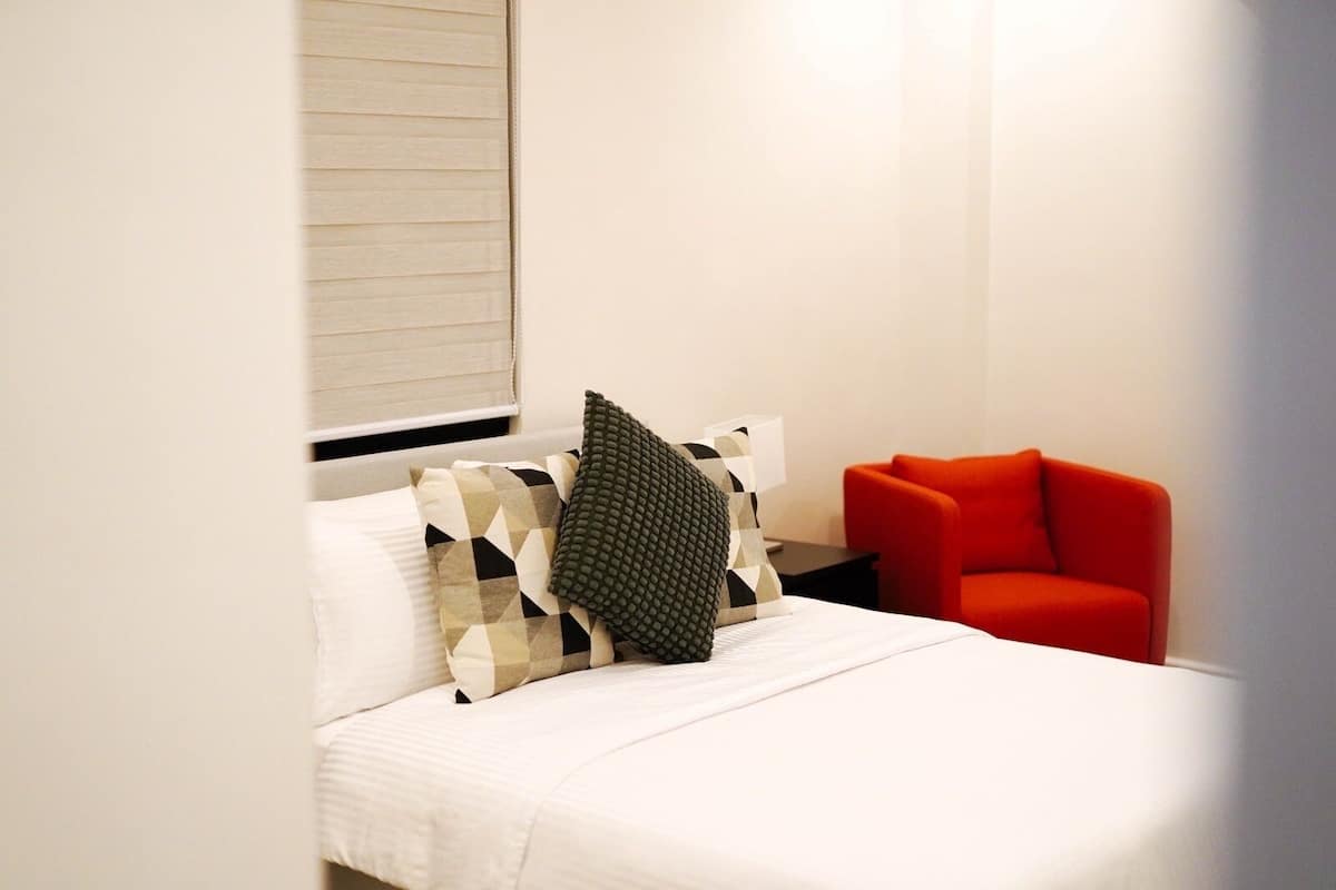 A hotel room with a bed, a nightstand, a red chair, and a window with blinds.