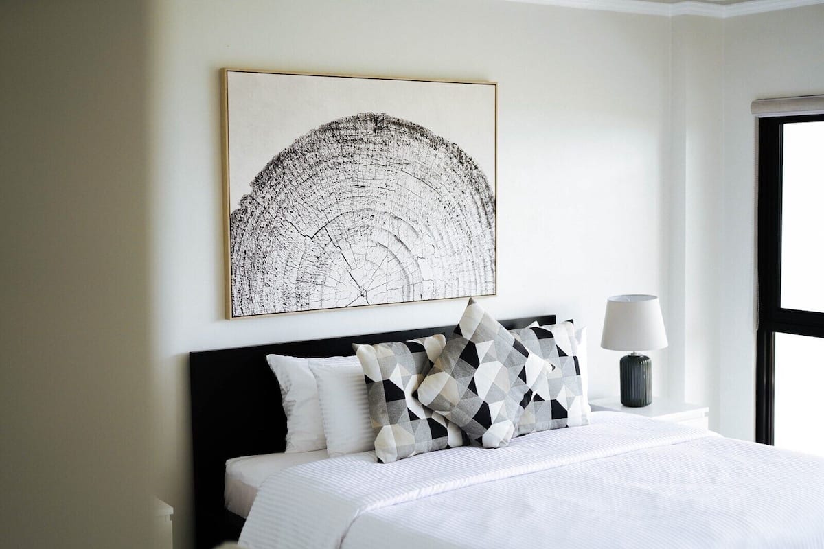 A neatly made bed with white bedding and patterned pillows, a bedside lamp, and a large abstract artwork on the wall.
