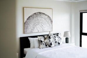 A neatly made bed with white bedding and patterned pillows, a bedside lamp, and a large abstract artwork on the wall.