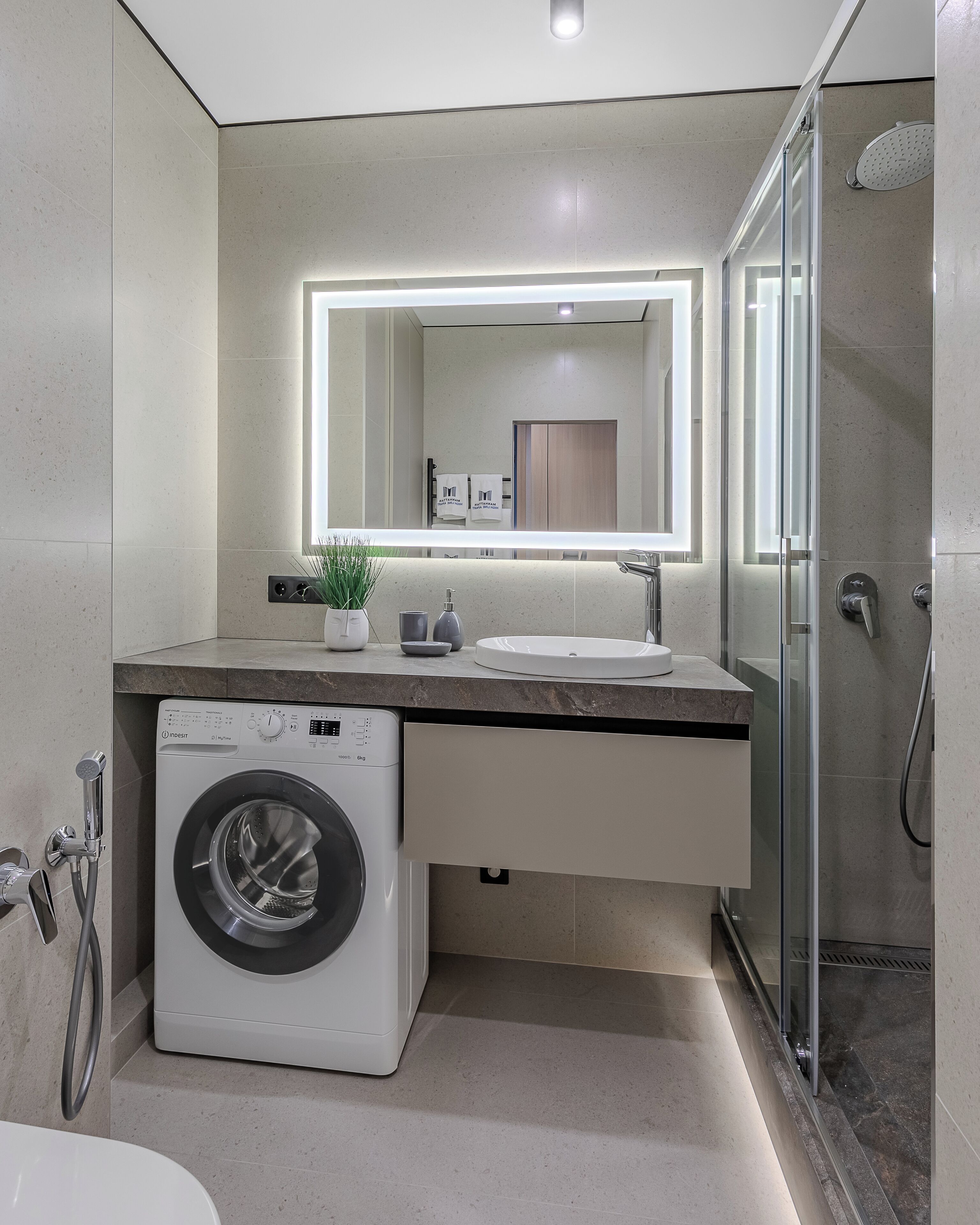 A modern bathroom with a built-in washing machine, a glass shower enclosure, and a large mirror with integrated lighting.
