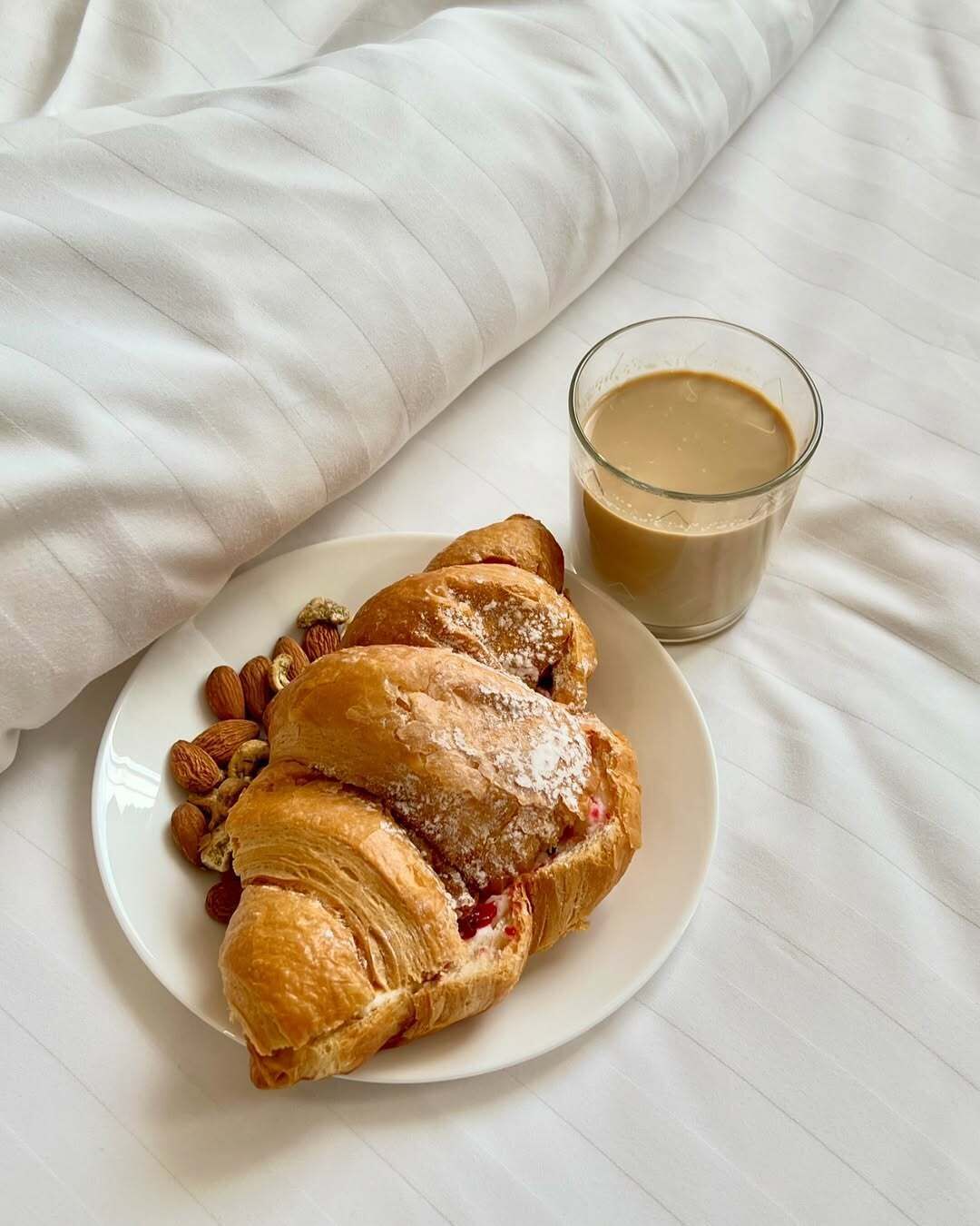 Um prato com um croissant e amêndoas, e um copo de café sobre um lençol branco.