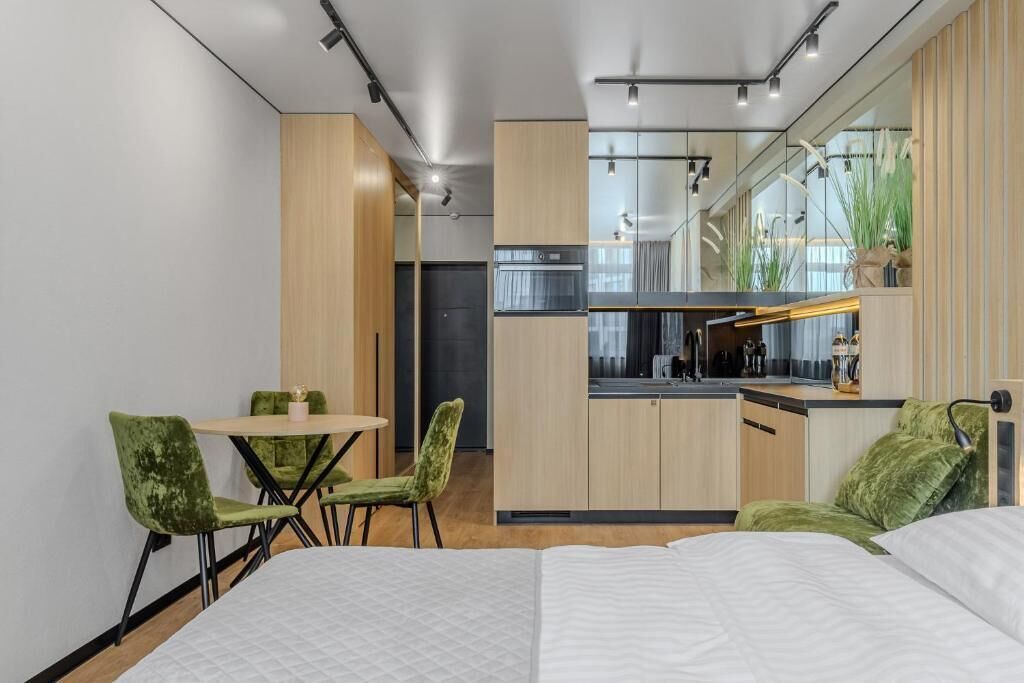 A modern hotel room with a bed, a dining area with chairs, a kitchen with a stove and sink, and a large mirror.