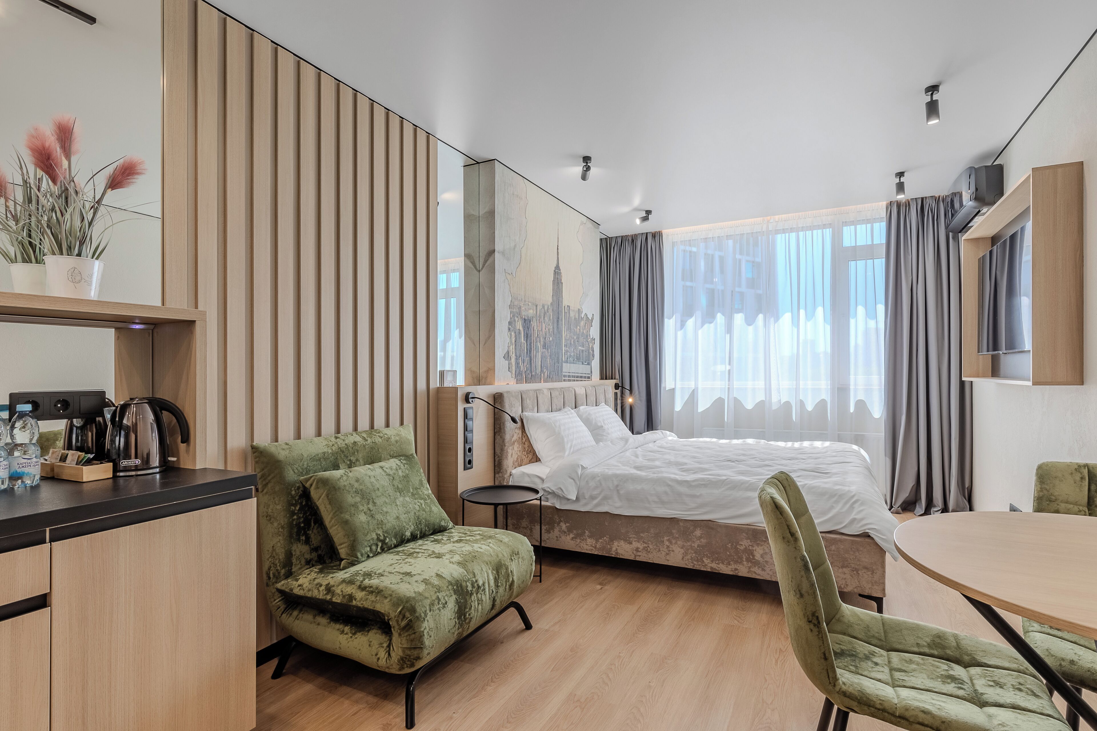 Een moderne hotelkamer met een bed, een groene fauteuil, een klein tafeltje en een kitchenette met een waterkoker.