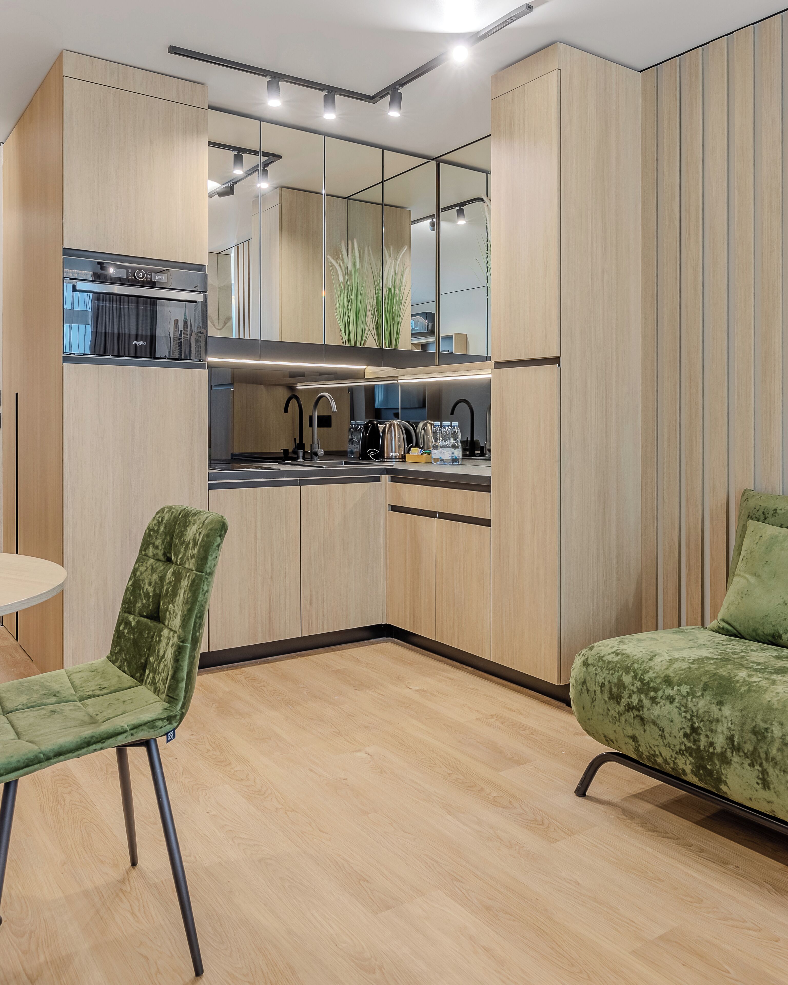 A modern kitchen with wooden cabinets, a built-in oven, and a mirrored backsplash. There is a green upholstered chair and a small round table.