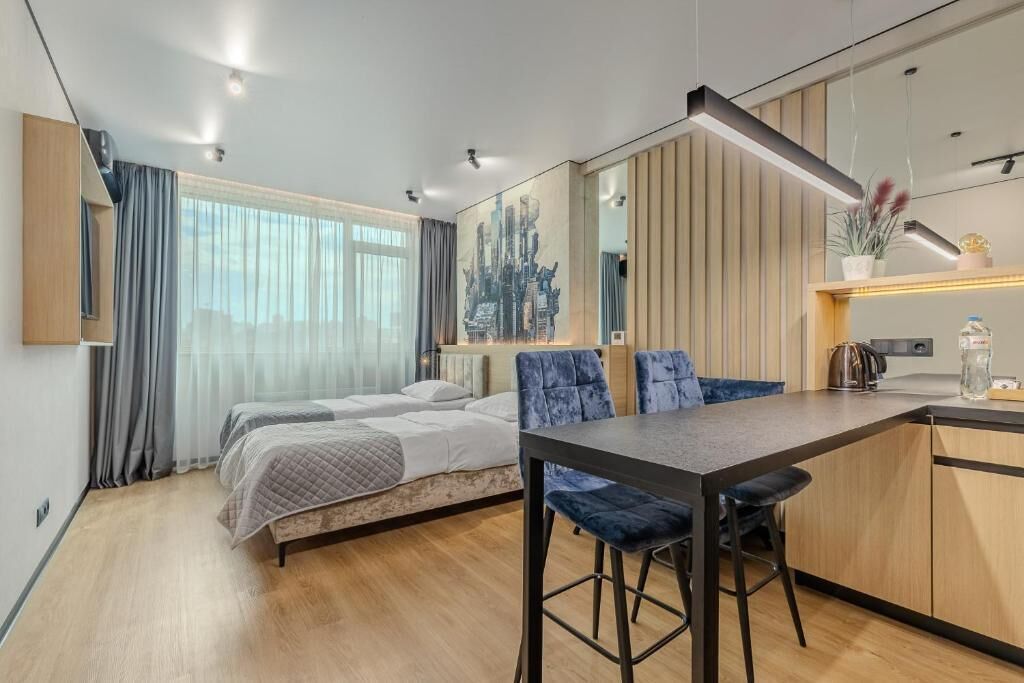 A modern hotel room with a bed, a small kitchenette, a dining area with a table and chairs, and a large window with curtains.