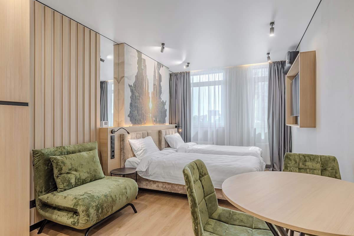 Ein modernes Hotelzimmer mit einem großen Bett, zwei grünen Polsterstühlen, einem runden Holztisch und einem Stadtbild an der Wand.