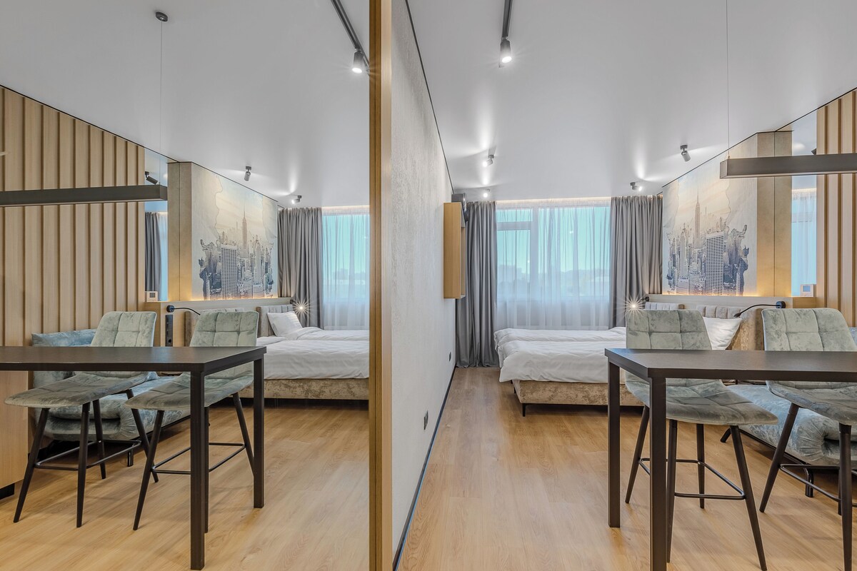 Habitación de hotel moderna con dos camas, una mesa de comedor y un mural con vista a la ciudad en la pared.