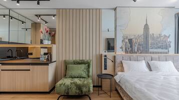 A modern hotel room with a wooden floor, a green armchair, a small round table, a bed with white linens, a cityscape mural, and a kitchenette with a sink and countertop.