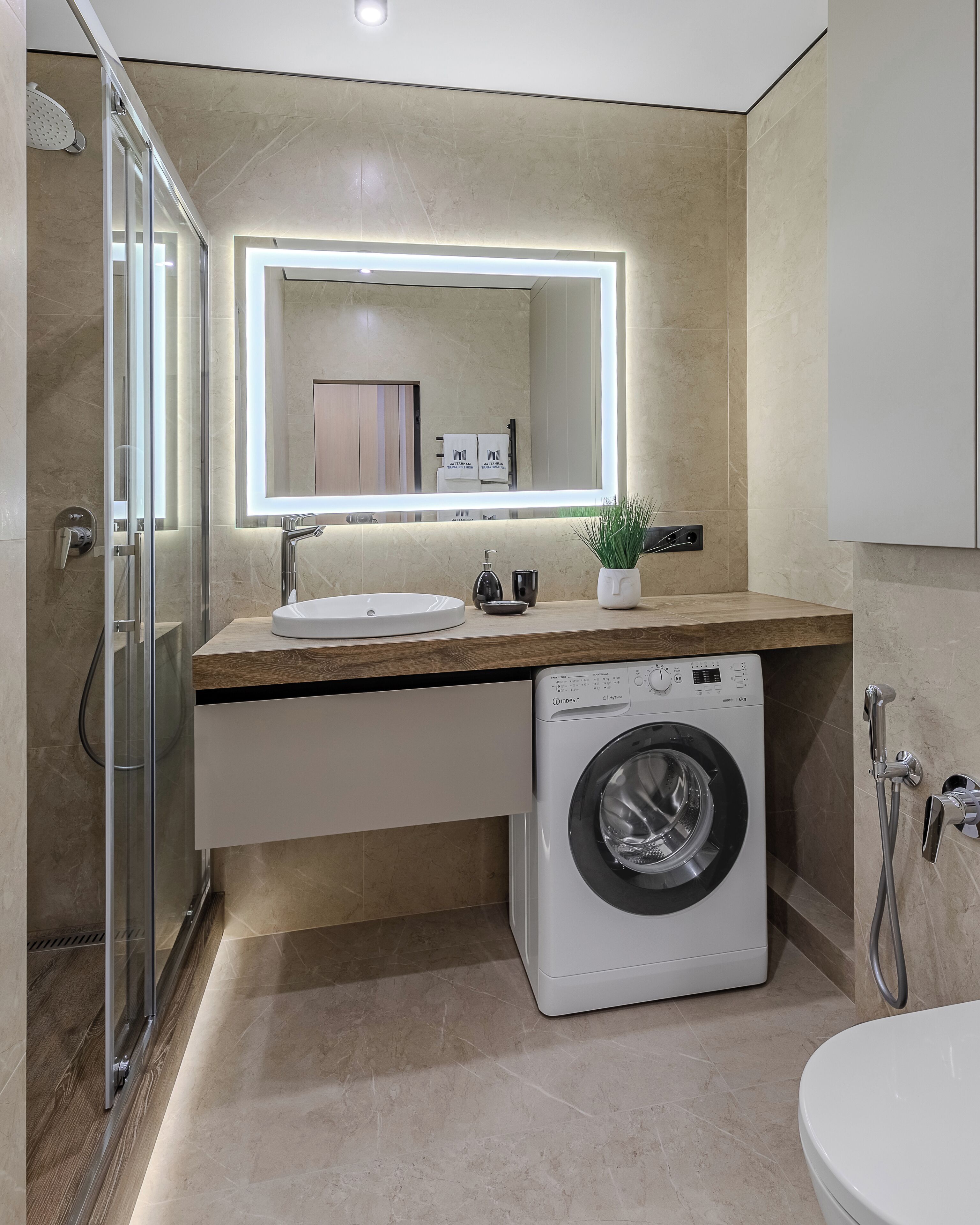 A modern bathroom with a glass shower enclosure, a built-in washing machine, and a large mirror above the sink.