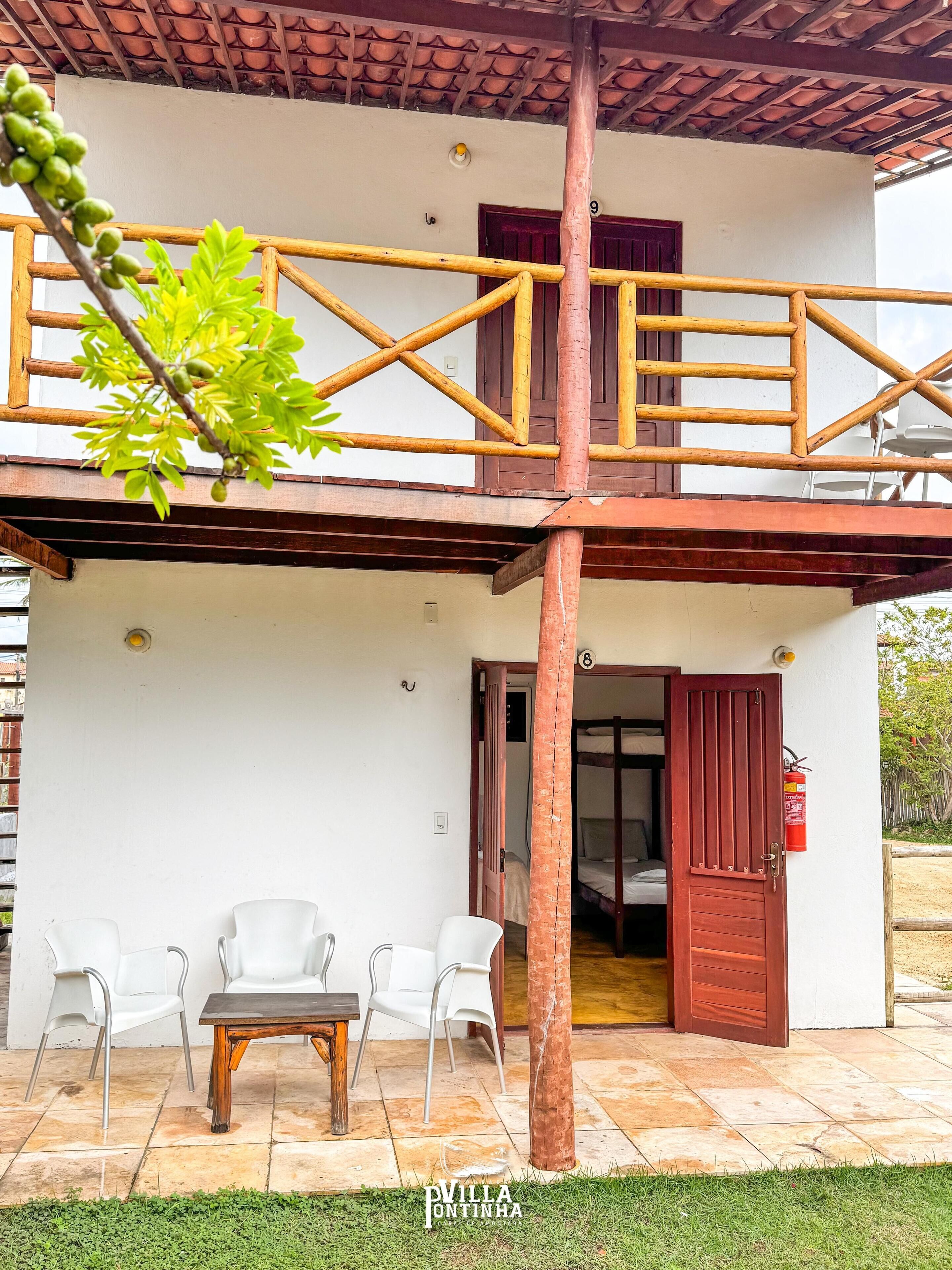 A two-story building with a balcony, a wooden table, and white chairs in the courtyard.