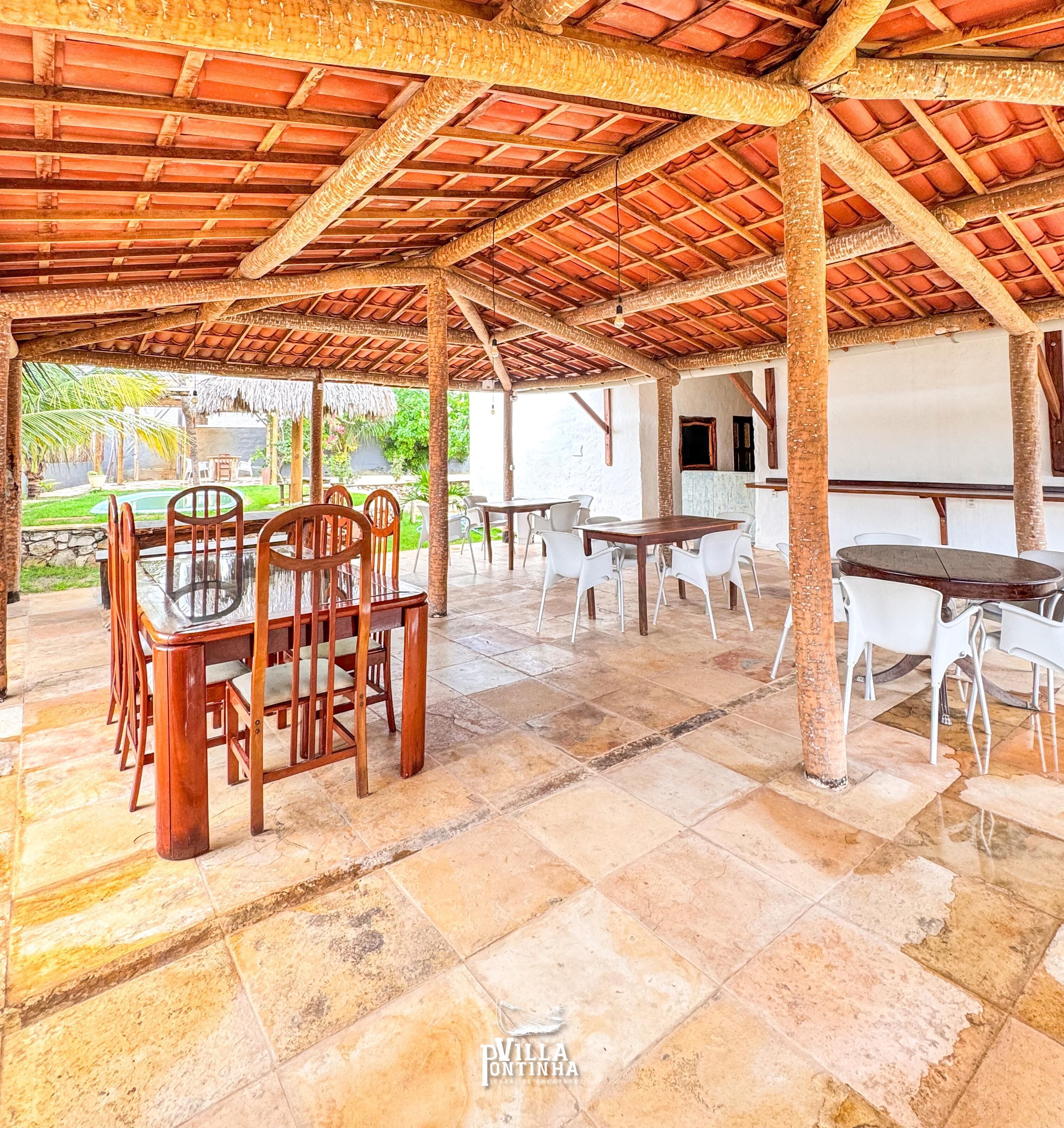 Um pátio coberto ao ar livre com mesas e cadeiras de madeira, piso de pedra e vista para uma piscina e vegetação.