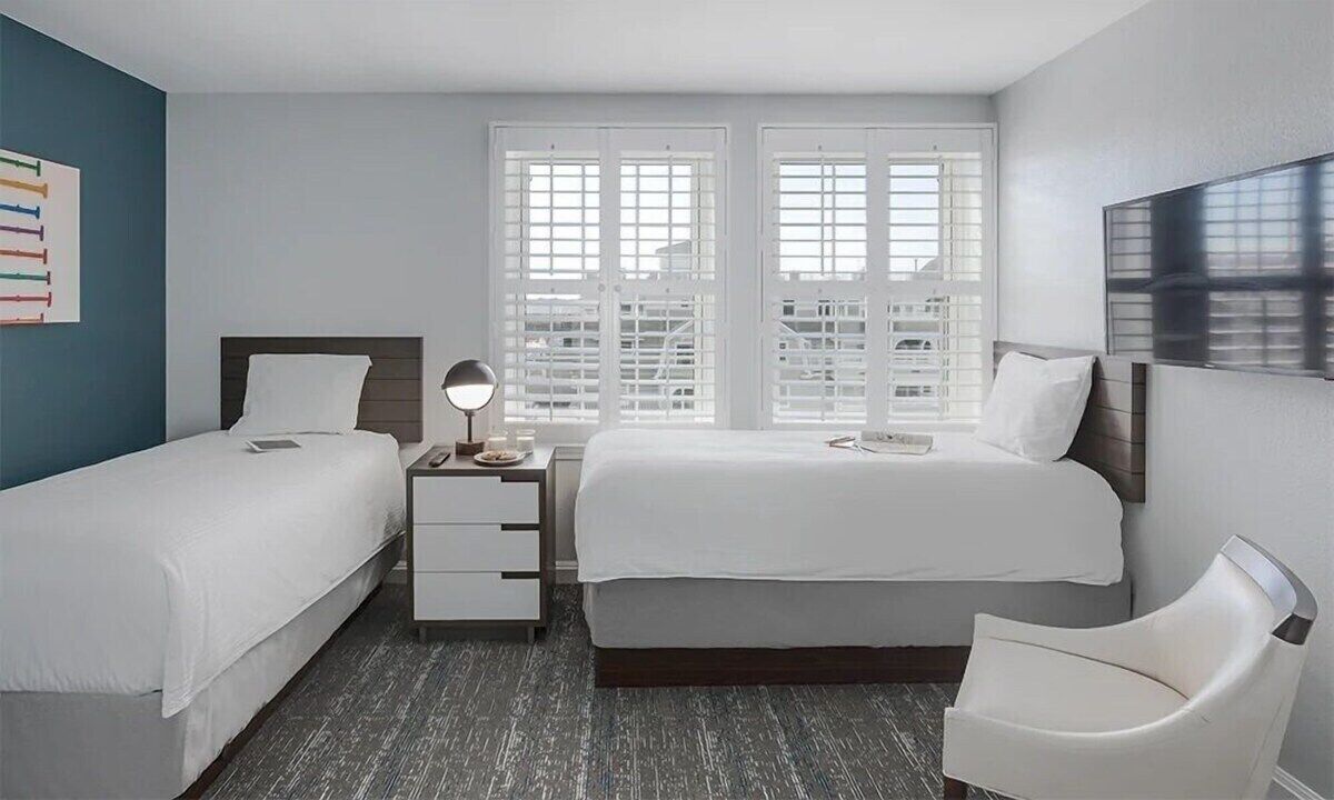 A hotel room with two beds, a nightstand, a lamp, and a chair.