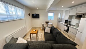 Una cocina moderna con armarios blancos, un microondas y un refrigerador. Hay un comedor con una mesa y sillas, y un sofá negro en la sala.