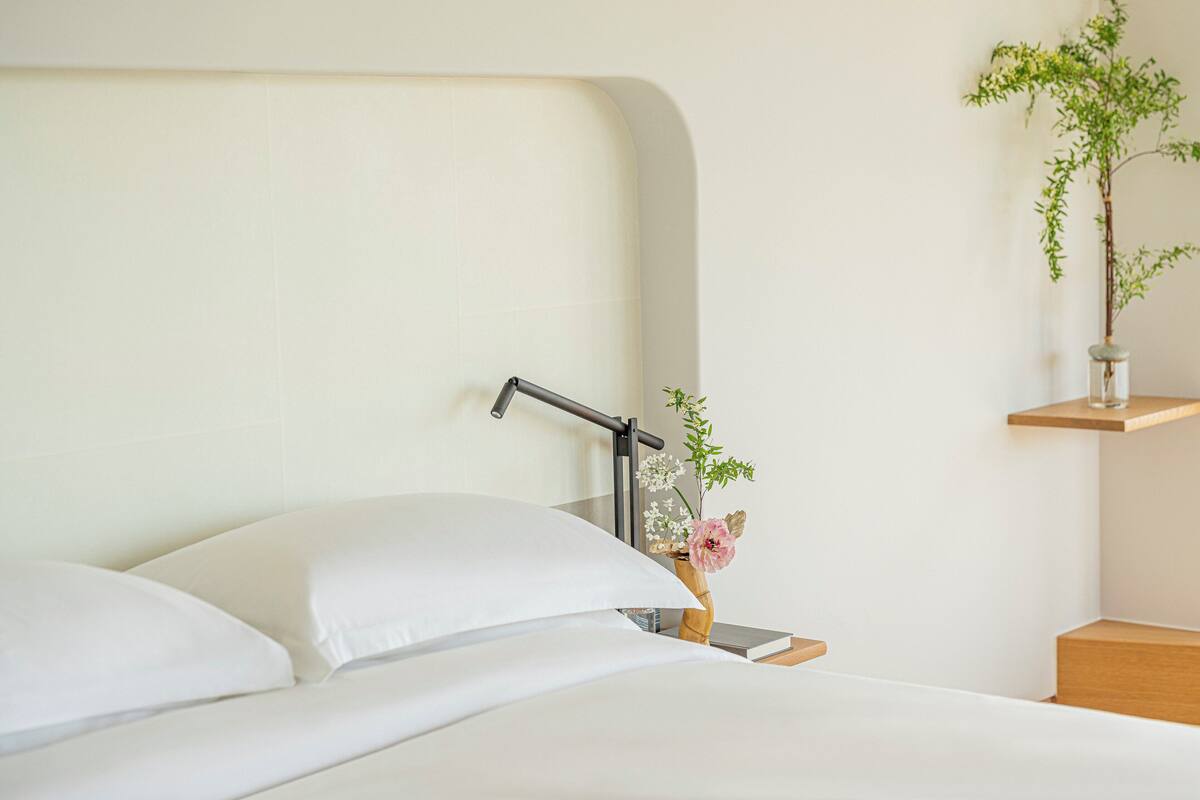 Een keurig opgemaakt bed met witte lakens, een nachtkastje met een smartphone, een kleine vaas met bloemen en een wandplank met een plantje.
