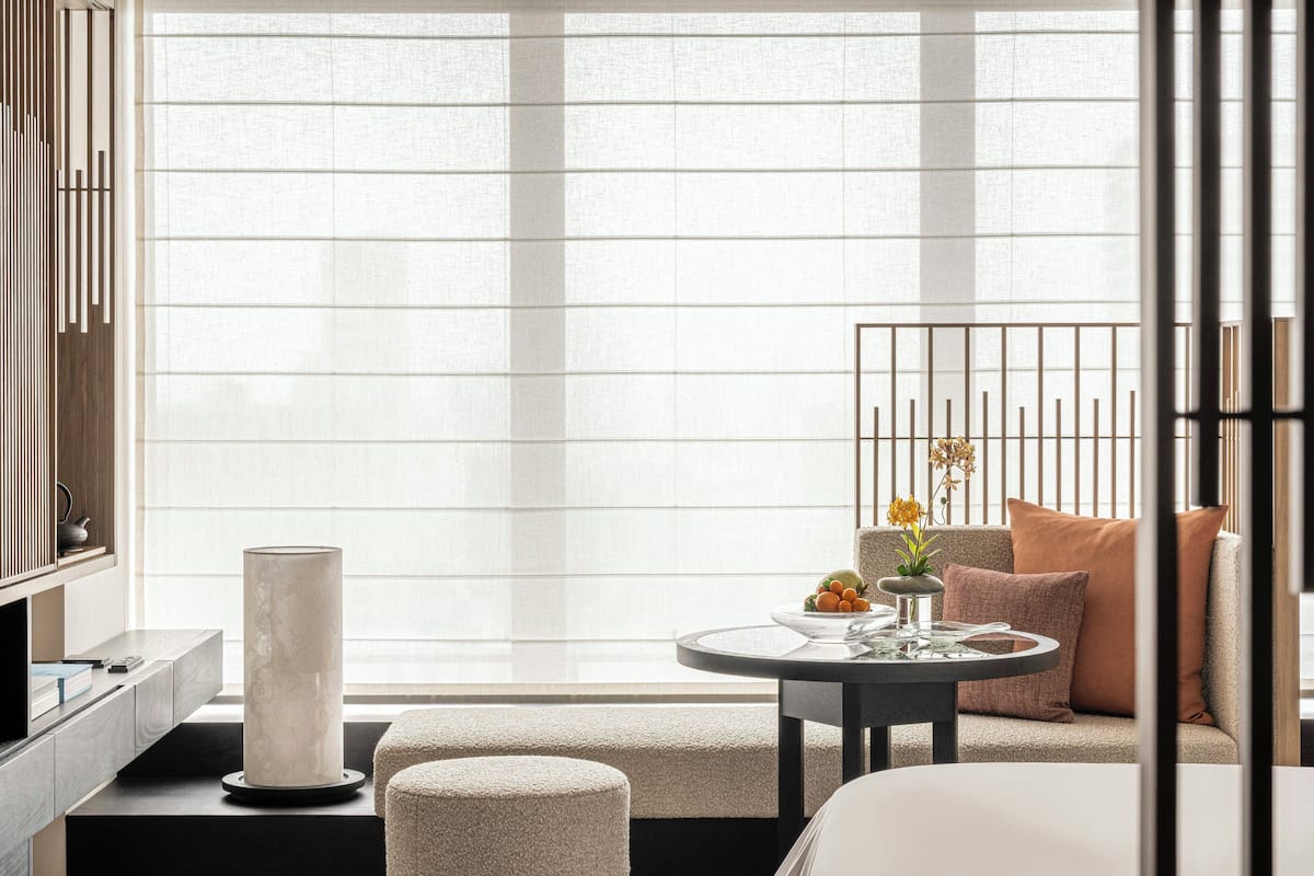 Een moderne hotelkamer met een groot raam, een ronde tafel met een bloevas, een bank met kussens en een hoge, cilindervormige lamp.