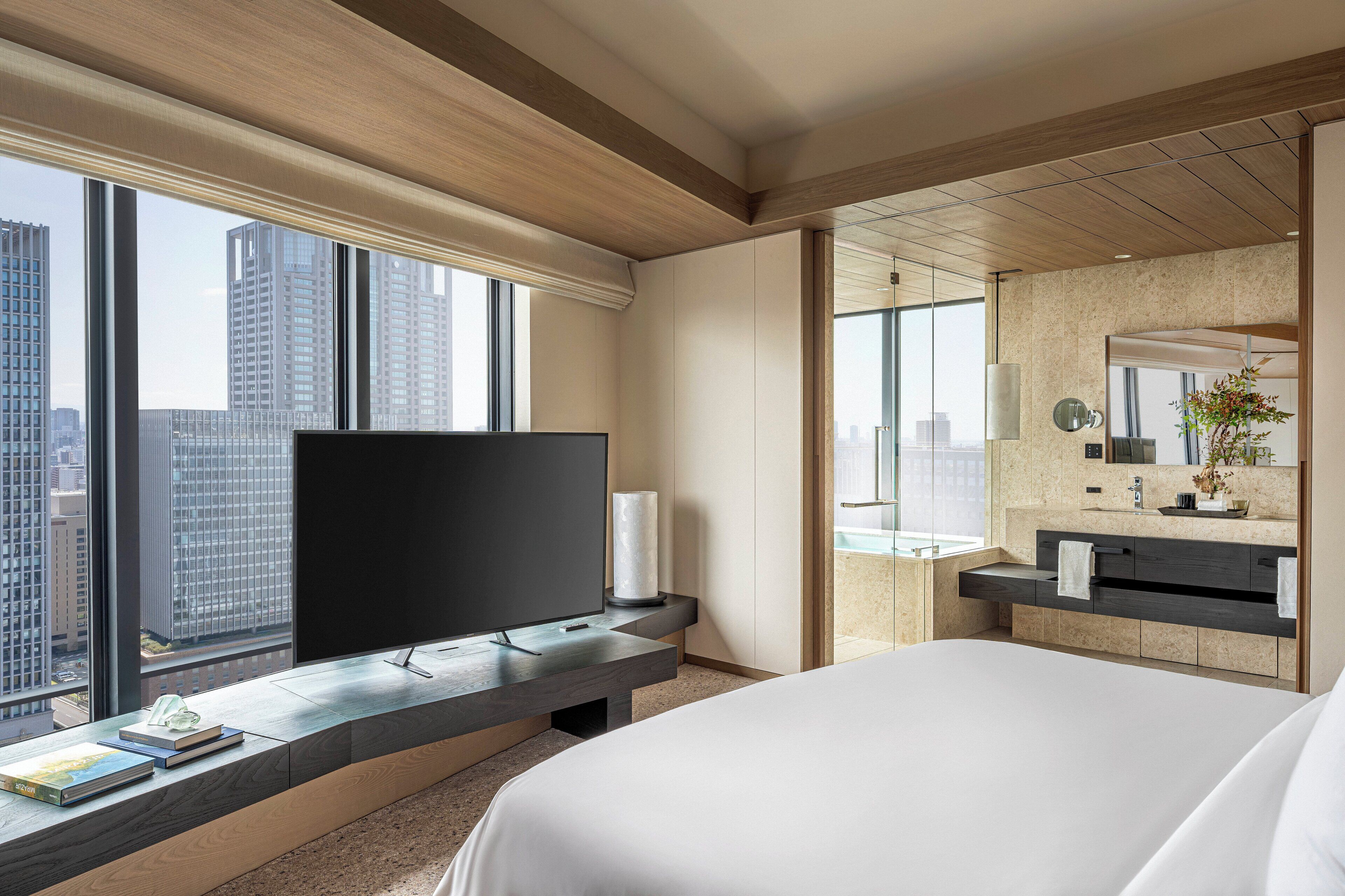 A modern hotel room with a large bed, a flat-screen TV on a wooden console, a bathroom with a large mirror and a sink, and a city view through large windows.