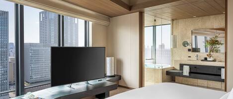 A modern hotel room with a large bed, a flat-screen TV on a wooden console, a bathroom with a large mirror and a sink, and a city view through large windows.